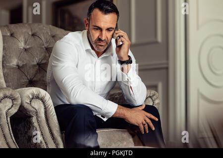 Geschäftsmann einen Anruf nach der Ankunft im Hotelzimmer. Mann in formale Abnutzung sitzt auf einem Stuhl und sprechen auf dem Smartphone. Stockfoto