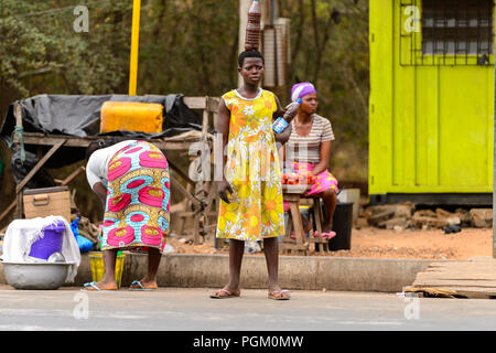 PIRA, BENIN - Jan 12, 2017: Unbekannter beninischen Frau im gelben Kleid mit Blumen hält eine Flasche mit Flüssigkeit. Benin Menschen leiden unter der Armut aufgrund Stockfoto