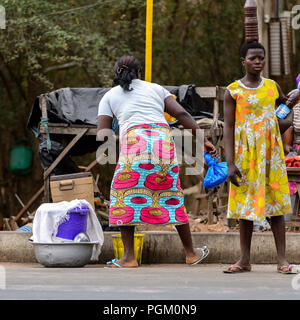 PIRA, BENIN - Jan 12, 2017: Unbekannter beninischen Frau im gelben Kleid mit Blumen hält eine Flasche mit Flüssigkeit. Benin Menschen leiden unter der Armut aufgrund Stockfoto