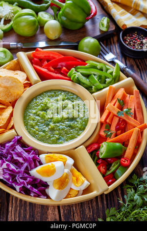 Grüne Salsa verde und mexikanischen Satz von gehackten frischen Gemüse, hartgekochte Eier und Pommes in Schalen auf alten rustikalen Holzbrett mit Zutaten, vertic Stockfoto