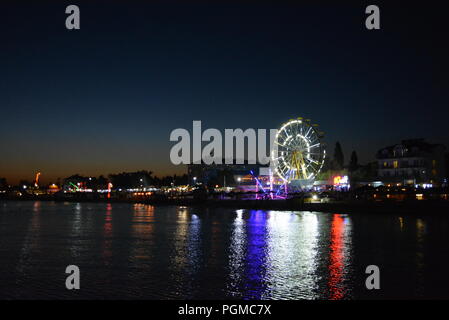 Nachtunterhaltung und Entspannung am Strand des Schwarzen Meeres mit Attraktionen, weißem Riesenrad, Trampolin in einer hellen, blauen, gelben Farbe. Stockfoto