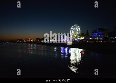 Nachtunterhaltung und Entspannung am Strand des Schwarzen Meeres mit Attraktionen, weißem Riesenrad, Trampolin in einer hellen, blauen, gelben Farbe. Stockfoto