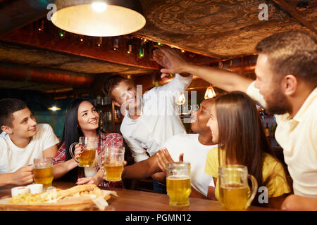 Gesellschaft von Freunden sprechen und trinken in der Bar. Einer der Männer, die gerade in der Kommenden und Gruß Jungs am Tisch, die hohe Fünf einer von Ihnen. Alle Menschen Stockfoto