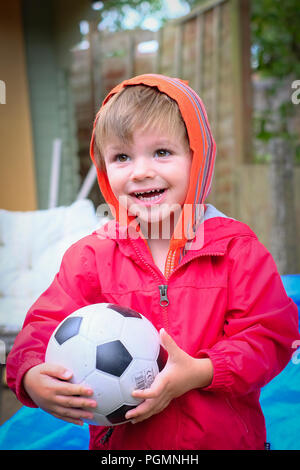 Jungen im Alter von zwei (2) mit roten Mantel und Fußball außerhalb glücklich und lächelnd. Stockfoto