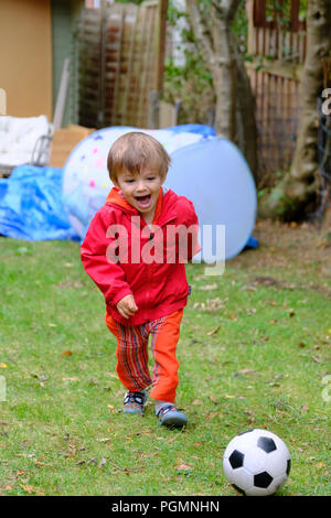 Jungen im Alter von zwei (2) mit roten Mantel und Fußball glücklich und lächelte, als er mit Fußball im Garten spielt. Stockfoto