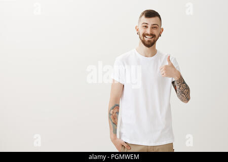 Portrait von good-looking Happy männliche Kursteilnehmer mit Bart und Tattoos, Daumen hoch und lächelte freundlich, Genehmigung geben, mögen neue Ideen während der Tagung in der Kunst Salon, stehend über graue Wand Stockfoto