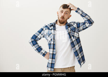 Lustig, gut aussehenden Kaukasier Männlich student mit Bart in Blue Plaid Shirt, die Vorbereitung auf die Prüfung, kratzen Kopf und schaute verwirrt und ratlos, keine Ahnung, was zu beantworten, über graue Wand in Frage gestellt Stockfoto