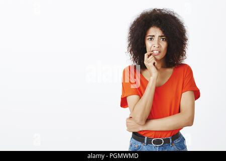 Mädchen Gefühl erschrocken und ängstlich, Anhörung schreckliche Nachrichten. Portrait von schockiert nervös Afrikanisch-amerikanische Frau mit Afro Frisur, sie beissen Nagel und runzelte die Stirn und starrte auf Kamera über graue Wand besorgt Stockfoto