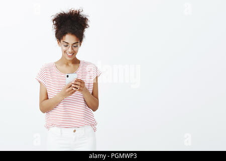 Studio shot von charmanten unbeschwerte junge Frau mit gebräunter Haut in Gläsern und Gestreiftes T-Shirt, suchen und lächelnd am Bildschirm des Smartphones, die angenehme Nachricht und beantworten über Gerät und WLAN Stockfoto