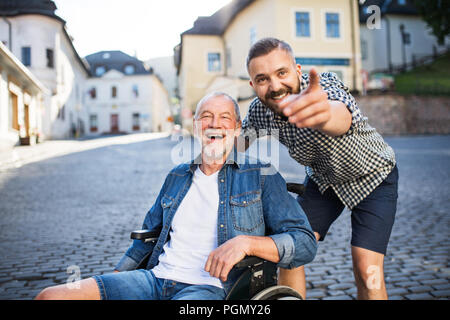 Ein erwachsener hipster Sohn mit seinem Vater im Rollstuhl auf einen Spaziergang in der Stadt und zeigt auf etwas. Stockfoto