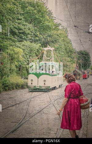 Crich Straßenbahn Dorf, 1940 s Wochenende. Gealtert - vintage street scene mit offenen Blick - top Straßenbahn in gepflasterten Straße unterwegs. Dame im roten Kleid wartet. Stockfoto