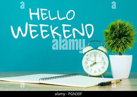 Hallo Wochenende Text auf grünem Hintergrund und kleinen Baum in weiße Vase mit Vintage Wecker und Notebook Papier Stift auf Holztisch. Stockfoto