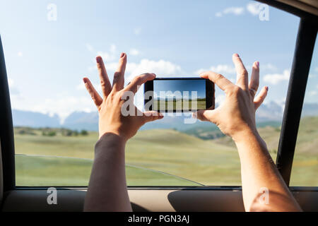 Nahaufnahme eines jungen Touristen im Auto macht ein Foto auf dem Telefon oben auf einem Berg mit Schnee vor einem blauen Himmel Hintergrund abgedeckt. Reisen Lifestyle und Stockfoto