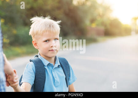 Vater an der Hand kleinen Sohn. Wenig Schüler mit Rucksack in die Kamera schaut und in die Schule zu gehen. Zurück zum Konzept der Schule. Stockfoto