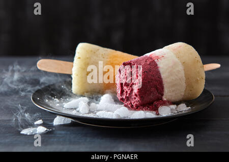 Obst Kaffee hausgemachtes Eis lolly auf einem Stick in eine schwarze Platte mit Scheiben von Eis auf einem Holz- dunkel Tabelle. Kaltes Dessert Stockfoto