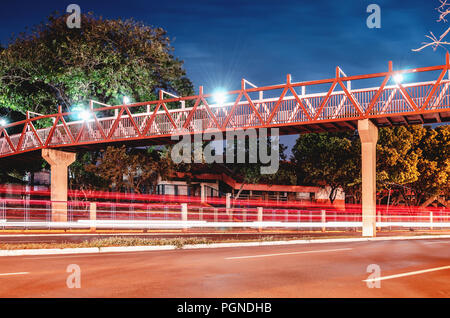 Rote Fußgängerbrücke über der Straße. Leichte Spuren von der vorbeifahrende Autos auf der Straße. Red Bridge bei Nacht Der horto Florestal in Campo Grande M Stockfoto