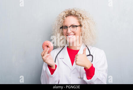 Junge blonde dietist Arzt Frau über Grunge graue Wand holding Donut zufrieden mit Lächeln tun ok Zeichen, Daumen hoch, mit den Fingern, ausgezeichnete Zeichen Stockfoto