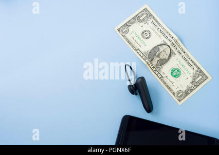 Geld Banknote mit schwarz Wireless Headset und Schwarz Handy am Schreibtisch mit Pastellfarben hellblau Hintergrund platziert. Flach, Platz für Text. Telekommunikation Stockfoto