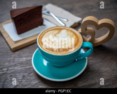 Kaffee latte Art in blau Tasse mit einem Stück Schokolade Kuchen, Gabeln und hölzerne Herz auf hölzernen Tisch Hintergrund. Kaffeeliebhaber Konzept. Stockfoto