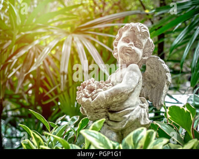 Alte vintage Cupid statue halten Sie die Blumen im Korb auf grüne Natur Hintergrund mit Sonnenlicht. Stockfoto