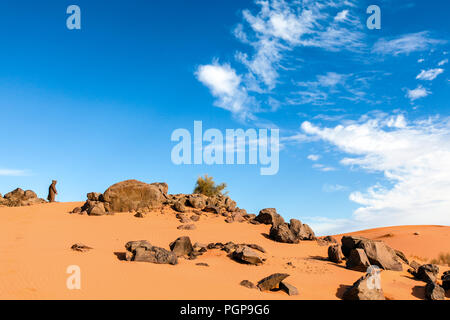 Erg Chebbi, Marokko. Alte Mann mit einem djellaba Robe steht durch einige Felsbrocken in den Sand und in die Ferne schaut. Blue Sky. Platz kopieren Stockfoto