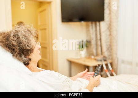 Ältere Frau im Bett in Ihrem älteren Apartment liegen mit Fernseher im Hintergrund Stockfoto