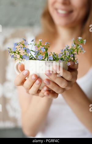 Junge Mädchen in einem weißen Hemd Holding eine gepunktete Tasse mit Vergißmeinnicht. Blumen in einem Becher. Hände mit Blumen. Lächelnde Frau mit Blumen. Tenderne Stockfoto