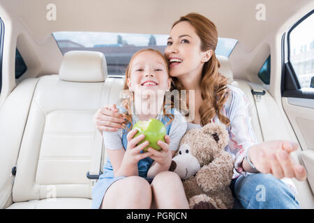 Portrait der glücklichen Mutter und Tochter mit Teddybär und Apple im Auto sitzen Stockfoto