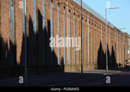 Silhouette eines Mannes, der entlang der Michaelson Road, Barrow in Furness, England, geht. Er wird von Mauern und Fenstern des BAE-Fabrikgebäudes in den Schatten gestellt. Stockfoto