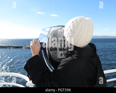 Ein junges Mädchen, für den Winter gekleidet, sieht durch ein Fernglas auf das Meer vom Deck eines Boots aus. Stockfoto