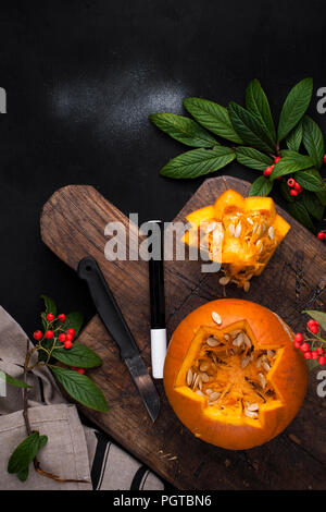 Gelber schöner Kürbis mit ausgeschnittendem Muster für den Herbsturlaub. Dekor für Halloween. Schwarzer Hintergrund und Holzbrett mit Beeren und Obst. Draufsicht. Hände in den Rahmen Stockfoto