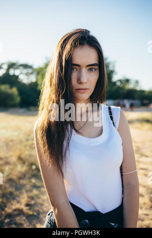 Close-up ehrliches Porträt der Schwere junge Frau mit langen braunen Haaren auf dem Hintergrund der Sommerwiese scheint die Sonne von hinten, vertikale Framing. Stockfoto