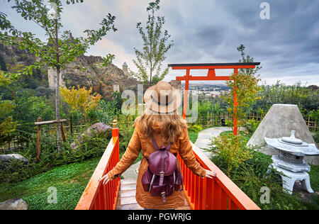 Frau in braunen Hut im japanischen Garten und das alte Schloss Narikala an bedeckt bewölkter Himmel im Botanischen Garten von Tiflis, Georgien Stockfoto