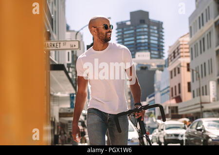 Hübscher junger Mann in Casuals auf der Straße mit dem Fahrrad. Afrikanischer Mann in casuals Walking im Freien in der Stadt mit dem Fahrrad. Stockfoto
