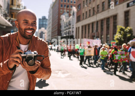 Lächelnd afrikanische Fotograf nimmt Bilder von Aktivistinnen auf der Straße. Mann Fotografieren von Menschen zu Fuß bei einer Rallye auf Straße der Stadt. Stockfoto