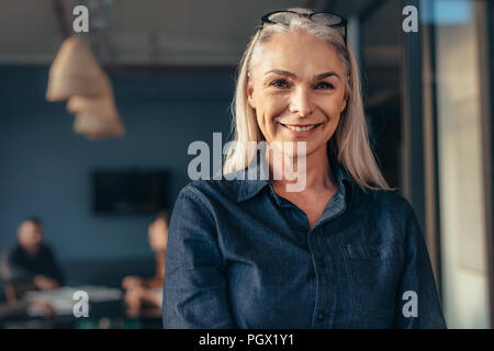 Nahaufnahme von Senior Business Frau im Amt auf Kamera und lächelnd. Reife Frauen im Amt mit dem Team im Hintergrund. Stockfoto