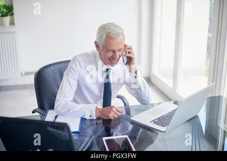 High Angle Shot der älteren Geschäftsmann mit Hemd und Krawatte, während im Büro Schreibtisch vor dem Laptop sitzt und spricht mit jemand auf seinem mobilen Ph Stockfoto