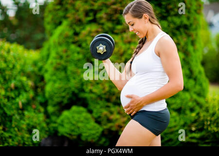 Die werdende Mutter Bauch berührt beim Anheben der Hantel in Park Stockfoto