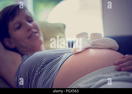 Weiß Baby Socken auf Bauch von Happy mittleren Alters schwangere Frau beim Entspannen. Stockfoto