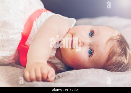 Portrait von cute adorable blonde Kaukasischen smiling baby Kind Mädchen mit blauen Augen in weißem Kleid mit roten Bogen liegend auf dem Bett und schaut träumend, Fee Stockfoto