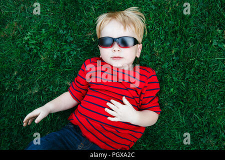 Close up Portrait von funny cute adorable White kaukasischen Kleinkind Kind Junge mit blonden Haaren im roten Pullover Sonnenbrille liegen auf grünem Gras. Anzeigen von