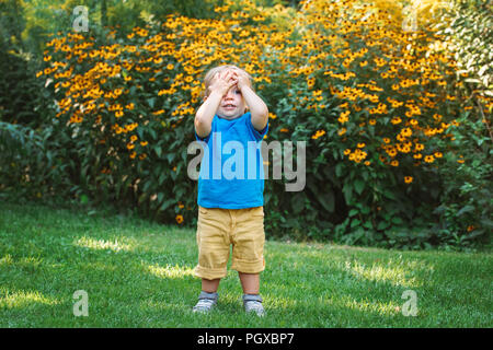 Portrait von cute adorable White kaukasische Baby Junge Kind ansehen unter gelben Blumen draußen im Garten Park seinen Kopf mit den Händen die Augen geschlossen halten Stockfoto