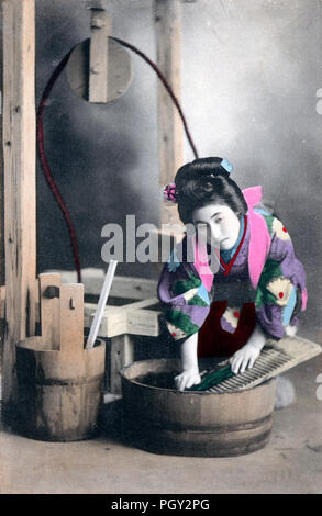 [1910s Japan - Japanische Frau im Kimono Wäsche] - Ein studio Foto einer jungen japanischen Frau im Kimono und traditionellen Kopfbedeckungen posiert als Hausfrau die Wäsche mit einem waschbrett und ein Oke (hölzerne Schaufel). 20. jahrhundert alte Ansichtskarte. Stockfoto
