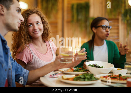 Lächelnde Mädchen mit gewellten Haar an ihrem Freund suchen während des Gesprächs durch serviert Tisch beim Abendessen mit Freunden Stockfoto