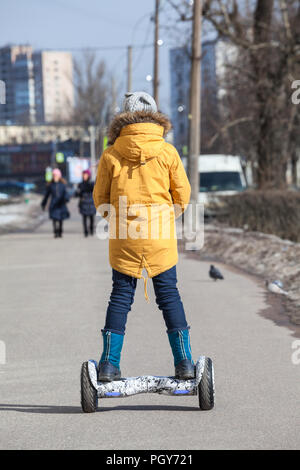 Ansicht von hinten an Mädchen in der gelben Jacke fahren auf Selbst-motiviert gyroscooter auf städtischen Straßen Stockfoto
