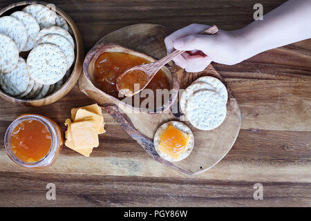Overhead shot der Hand einer Frau vorbereiten Wasser Cracker, Cheddar Käse und hausgemachten herzhaften Gesalzen Vanille Kantalupe Konfitüre Vorspeisen. Stockfoto