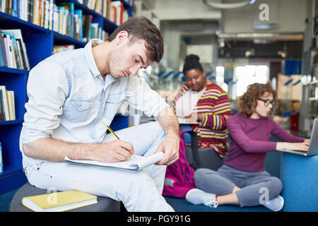 Männliche Kursteilnehmer schriftlich Bericht während der Sitzung in Bibliothek mit anderen multiethnischen Studenten im Hintergrund konzentriert Stockfoto