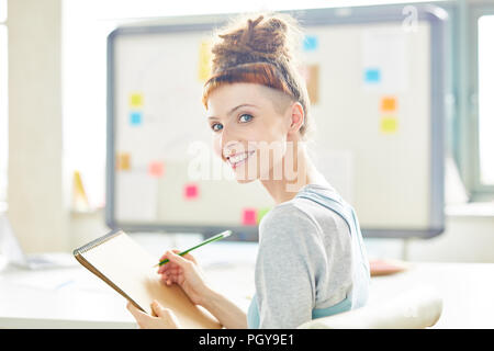 Fröhliche attraktiven weiblichen startupper mit dreads Schreiben in sketchpad und Kamera beim Sitzen auf den Vorsitz im Board Room Stockfoto
