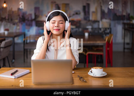 Schöne asiatische Frau entspannende und Hören von Musik im Cafe mit Laptop und Kaffee Tasse. Die Menschen und Lebensstile Konzept. Freiberufliche glücklich arbeiten