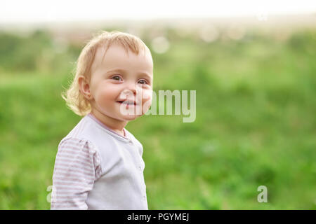 Lächelnd kleine Mädchen zu Fuß auf der grünen Wiese. Kind suchen Glücklich, positiv, tragen weiße Hemd mit rosa Streifen, mit großen grünen Augen, kurze blonde Stockfoto
